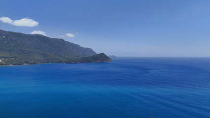 A breathtaking view of a vast blue ocean stretching to the horizon, bordered by a rugged, mountainous coastline under a clear, sunny sky. 