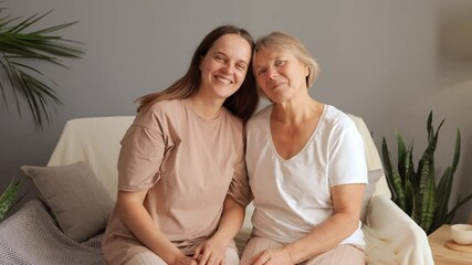 Elderly Caucasian woman and her adult daughter sitting together on couch smiling sharing joyful moment together at home hugging with love and looking at each other