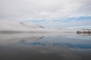 Fototapeta premium Spiegelnder See am Morgen mit reflektierenden Wolken und Nebel.