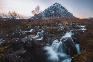 A breathtaking view of Buachaille Etive Mor and River Coupall waterfall in Glencoe, with snowcapped mountain peak and rugged winter landscape at sunset or sunrise in the Scottish Highlands, Scotland.