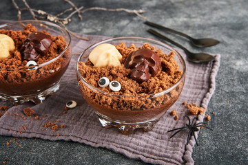 Halloween party chocolate puddings or Panna Cotta with brownie crumbles and chocolate sculls served in clear glass on grey dark background. Party idea for Halloween.