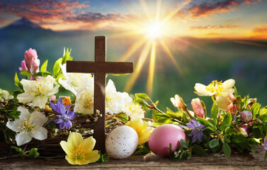 Small wooden Christian cross with flowers and easter eggs and mountain sunrise. 