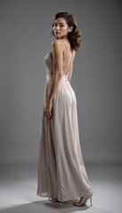 Graceful young woman in elegant gown posing with back turned against gray background