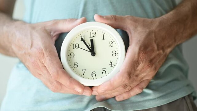 Health clock, adult man holding white alarm clock over his abdomen, 4K