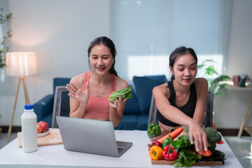 Two asian sportswomen greeting on a video call and preparing healthy food, discussing nutrition and fitness through a laptop in living room, wellbeing and healthy lifestyle concept