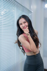 Asian woman wearing sportswear smiles and points her finger while standing next to a window with blinds, conveying a sense of playfulness and confidence
