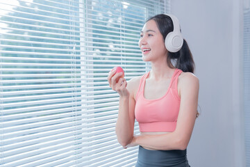 Happy asian sportswoman wearing headphones and eating apple while listening music near window with blinds at home, enjoying healthy lifestyle