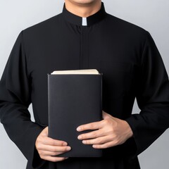 A priest in black cassock holds a closed book in his hands against a white background.