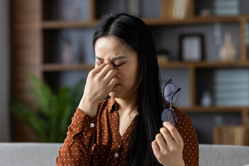 Asian woman with eye strain holding glasses, indoor stress and tiredness. Her expression reveals...
