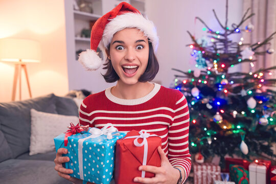 Excited young woman in Santa hat holding Christmas presents by decorated tree