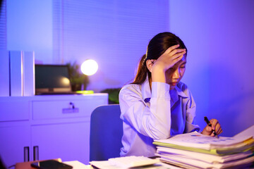 Asian woman diligently works overtime, managing a heavy workload with focus and determination....
