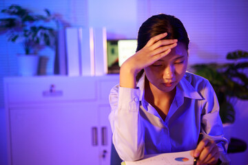 Asian woman diligently works overtime, managing a heavy workload with focus and determination....