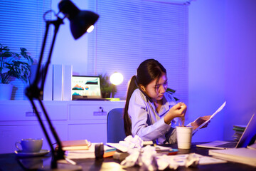 Asian woman diligently works overtime, managing a heavy workload with focus and determination....