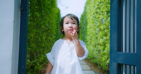 A cute Curious child girl in a white dress Exploring enjoys smiling playfully outdoors, standing posing in a lush green garden surrounded by greenery on a sunny day