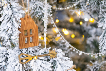 Naklejka premium Key and tiny house keychain in hand on background fairy lights Christmas tree. gift for New Year, Christmas. Building, design, project, moving, insurance, mortgage, rent and purchase real estate.