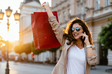 A smiling mid-adult Caucasian woman chats on her phone, holding up a red shopping bag, exuding...