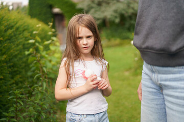 Little cheerful girl without one front tooth dressed in a T-shirt and jeans stands next to her mother in the backyard of her house in the summer. 