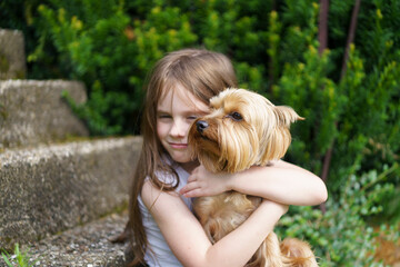 A little cheerful girl without one front tooth dressed in a T-shirt and jeans hugs her small dog sitting on the steps in the backyard of her house in the summer. 