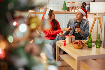 christmas, christmas eve, party, couple, xmas, celebration, decoration, giving, santa hat, enjoyment. A man and woman are sitting on a couch in a living room, holding wine glasses and smiling.