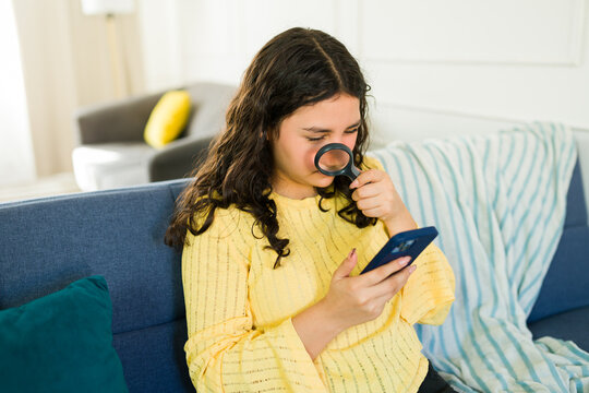 Female teenager with eyesight problems using a magnifying glass to look at her cellphone sitting on a sofa at home