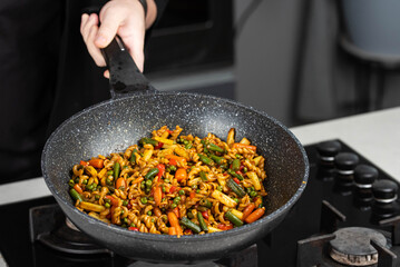 Close up of Chef cook hands cooking and toss roasted pasta, onion, pepper, carrot, vegetables, mushrooms, green peas, beans, garlic in frying wok pan on gas stove. Flying food levitation.