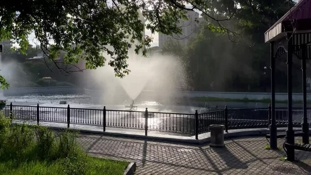 Beautiful fountain spraying water in city pond in summer with green trees around
