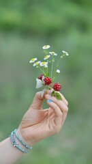 A hand with delicate bracelets holds a bouquet of small flowers and berries in a natural setting, evoking simplicity and appreciation for nature