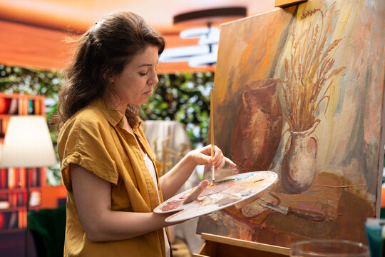 In a rustic workshop, artist mixes a variety of watercolor paint on a palette to create a colorful masterpiece on canvas. Woman painting with a brush and showing her skill, artwork creation.
