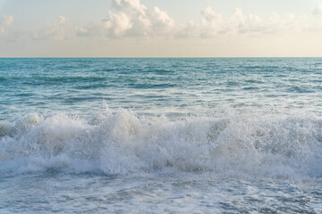 The sea waves roll onto a pebble sea shore against a blue sky with clouds.