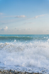 The sea waves roll onto a pebble sea shore against a blue sky with clouds.