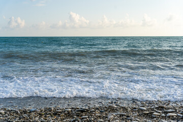The sea waves roll onto a pebble sea shore against a blue sky with clouds.