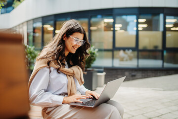 Fototapeta premium Young businesswoman working on laptop outside of modern business building