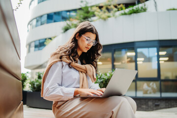 Young businesswoman working on laptop outside of modern business building