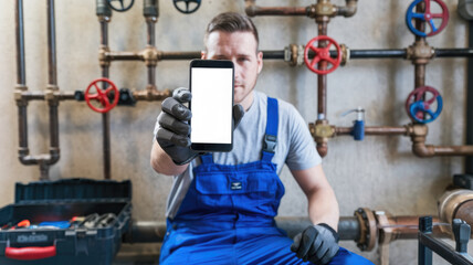 A man in blue overalls holding a cell phone with a white background. He is sitting on a bench