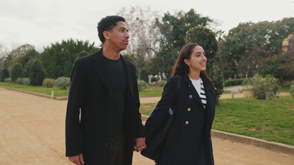 Young couple holding hands and walking in park with trees and grass on background