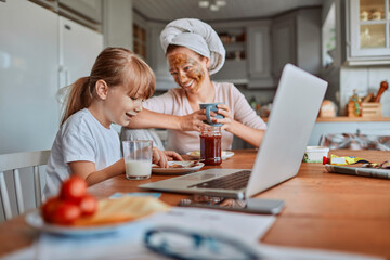 Mother and daughter having breakfast and watching laptop together in cozy kitchen