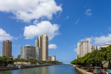 Naklejka premium The rippling waters of the Ala Wai Canal with skyscrapers, hotels and condos, lush green trees, cars on the street, blue sky and clouds in downtown Honolulu Hawaii USA