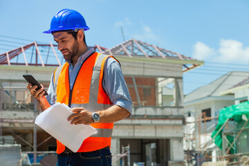 A man in a construction site is looking at his cell phone while wearing an orange vest. He is...