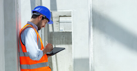 A man in a safety vest is using a tablet to take notes. He is wearing a blue hard hat and he is focused on his work