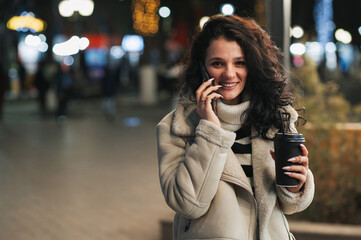 A smiling dark-haired girl stands outside on a winter evening with a paper cup of coffee in her hands and talks on the phone.