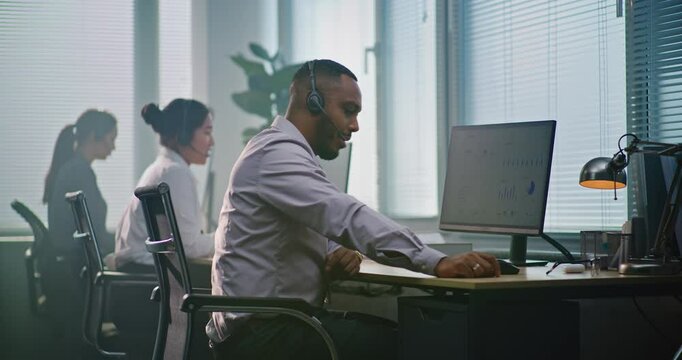 Multiethnic team of technical support operators working in modern call center office. African American helpdesk specialist talks on headset, uses computer, looks at camera providing customer service.
