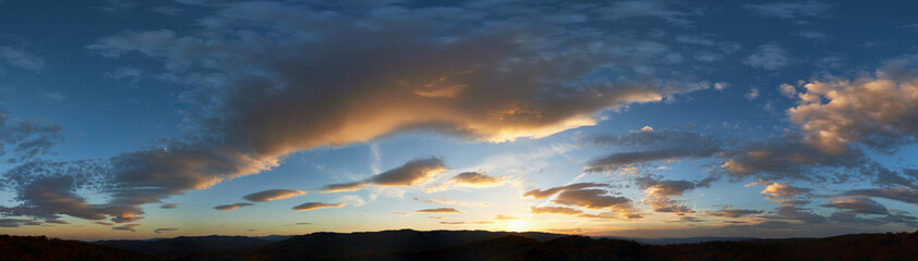 Stunning sunset fills sky with rich hues of blue and orange. Dramatic clouds with hints of pink and gold spread across sky. Silhouettes of hills in foreground add depth to breathtaking evening scene.