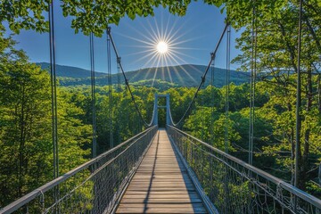 Obraz premium A scenic suspension bridge surrounded by lush greenery, with the sun shining brightly in a clear blue sky.