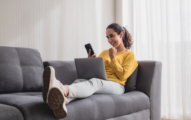 Obraz premium Business latin woman freelance one person smile cheeful happy sitting sofa and look laptop talk mobile phone plan marketing job work new project along with success future in living room home office.