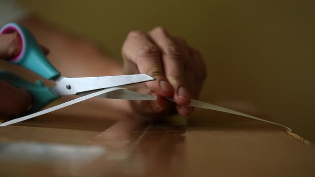 Close-up cropped shot of unrecognizable man carefully cutting plastic strap with scissors to open cardboard box, preparing to unpack delivered goods, showcasing meticulous unboxing process slow motion