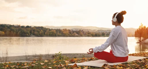 Fotobehang Lotusbloem Female person in headphones sits in lotus pose in front of autumn river in nature and meditating during sunset. Health lifestyle and city escape.  © Barillo_Images