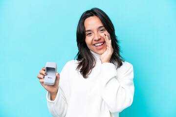 Fototapeta premium Young hispanic woman holding a engagement ring isolated on blue background shouting with mouth wide open