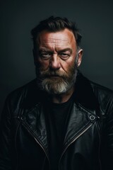 Obraz premium Intense lighting highlights a bearded man's features as he poses confidently in a black leather jacket. the dark, monochromatic background enhances the moodiness