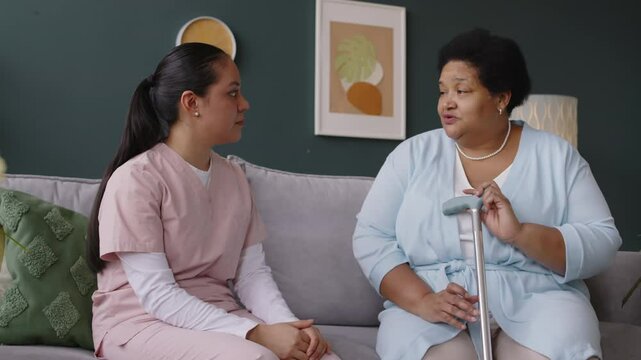 Hispanic young nurse listening carefully to Black elderly woman with cane talking about her health complaints while sitting on sofa in living room