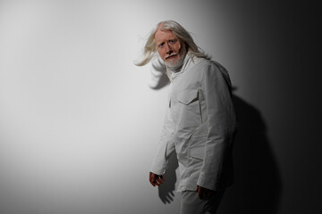 Elderly man with long white hair dressed in a white outfit, standing against a minimalist gray...